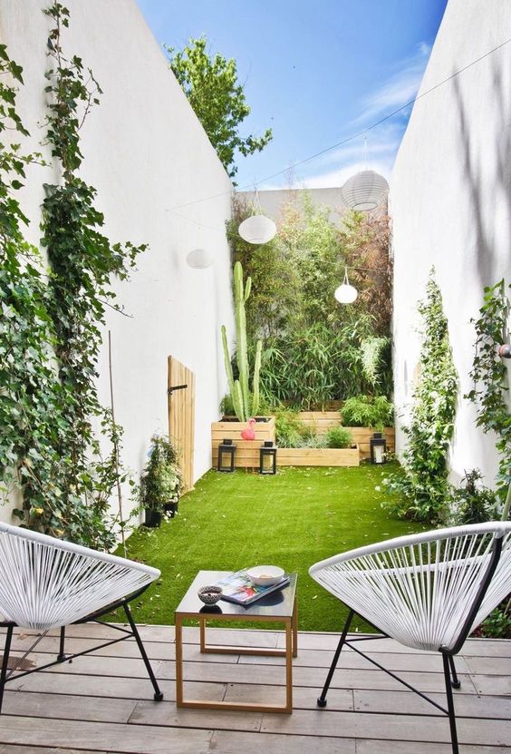 a small and fresh patio with a grene lawn and vines climbing up the walls, planked pots with plants, a deck with chairs and a table