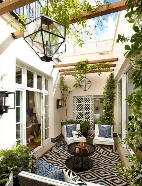 a small and welcoming patio with a rug, white chairs, a fire bowl, a sofa and lots of greenery and lamps