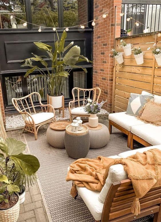 a small and welcoming patio with a sofa, a couple of chairs, poufs with blooms, lights and potted statement plants