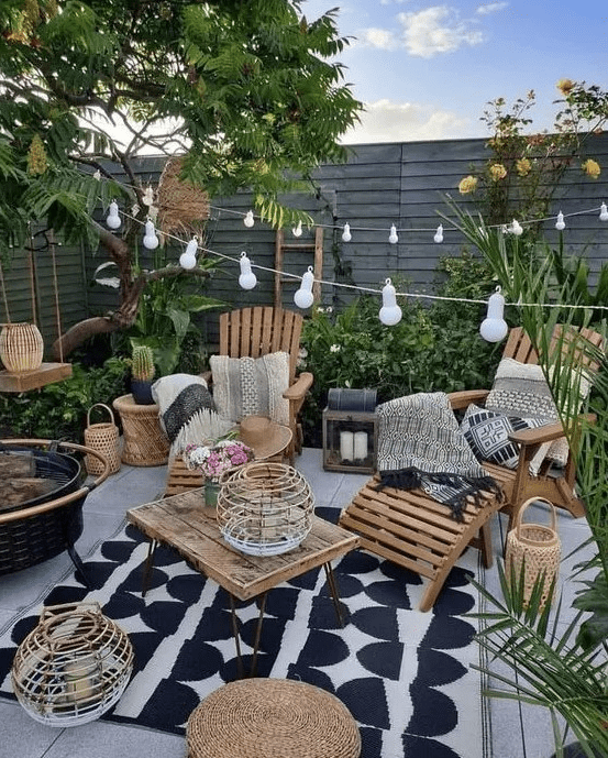 a small boho backyard with a wooden deck, wooden loungers, a table, some jute poufs, candle lanterns, string lights and greenery