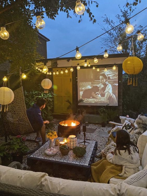 a small cinema patio with a fire bowl, a wicker sofa, a coffee table, a pendant chair and string lights over the space