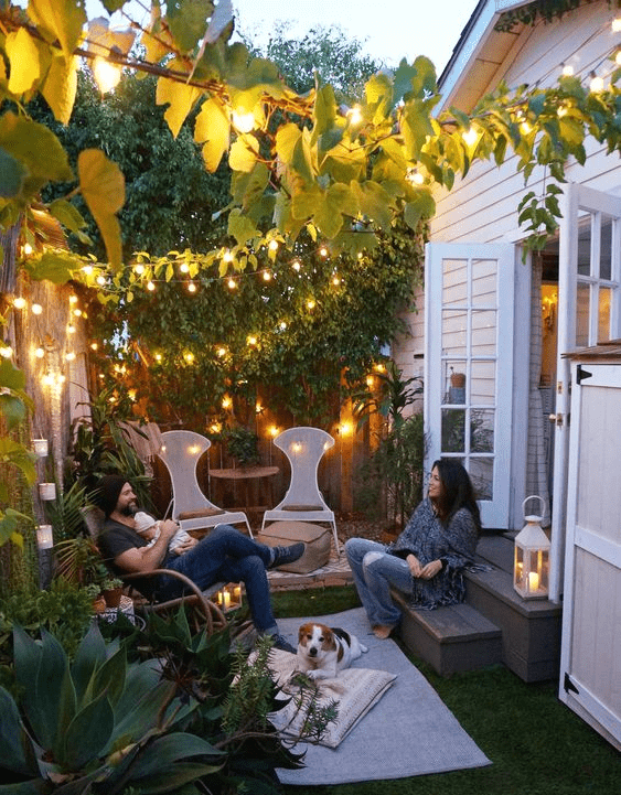 a small cozy patio with a couple of chairs and ottomans, some rugs and a pillow and lots of lights above