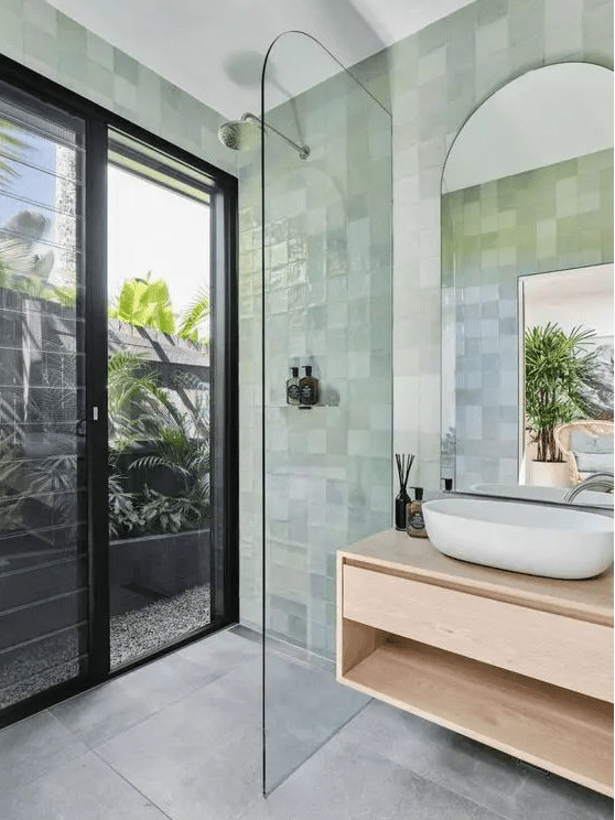 a small modern bathroom with a glass door that leads to the garden, green zellige tiles, a floating vanity and an arched mirror