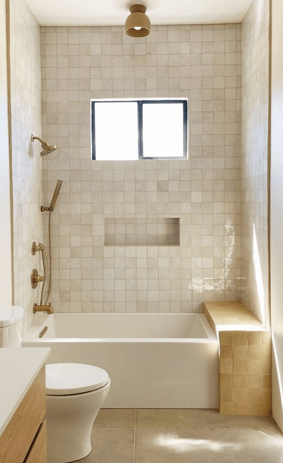 a small neutral bathroom clad with Zellige tiles, with a tub and a Zellige tile bench, brass fixtures and a window
