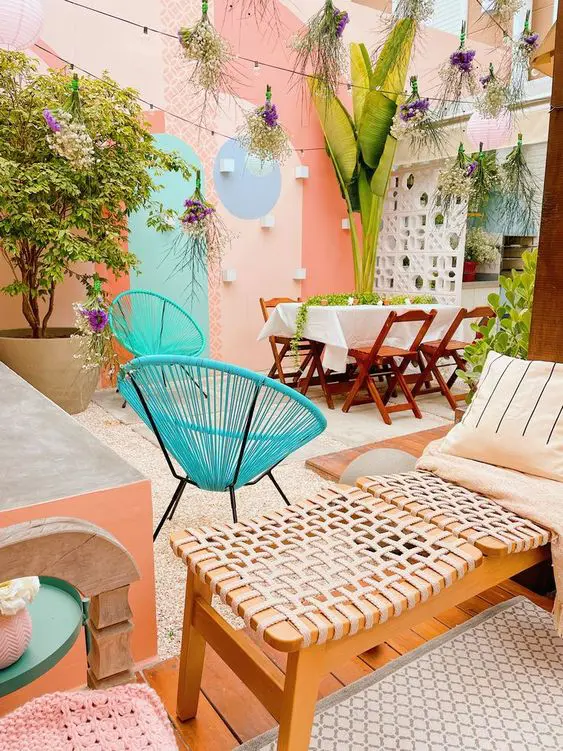 a small patio with peachy walls, a dining zone, potted plants and blooms, blue chairs, a bench and flowers hanging over the space