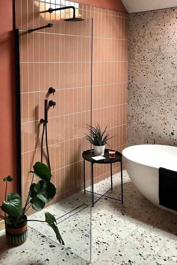 a stylish modern bathroom with a terracotta tile accent wall, grey terrazzo, an oval tub, a black side table and potted greenery