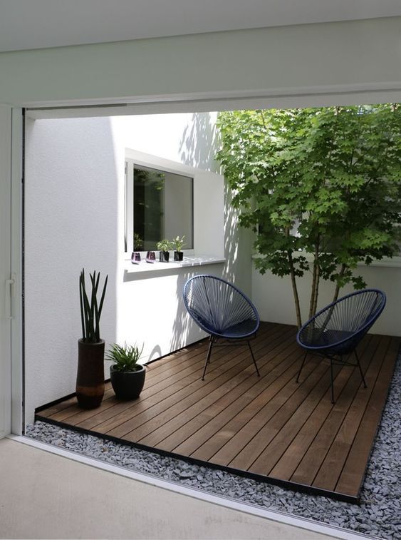 A tiny minimalist patio with a pass through window, a couple of chairs, a tree and potted plants, just a table is missing