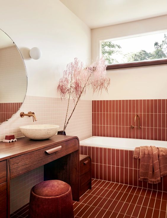 a warm earthy bathroom done with terracotta tiles, a tub, a stained vanity, a rust pouf and some pretty decor
