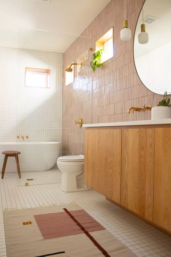 a welcoming bathroom with white square and pink Zellige tiles, a stained vanity, a shower space and a bathtub, a round mirror and a pendant lamp