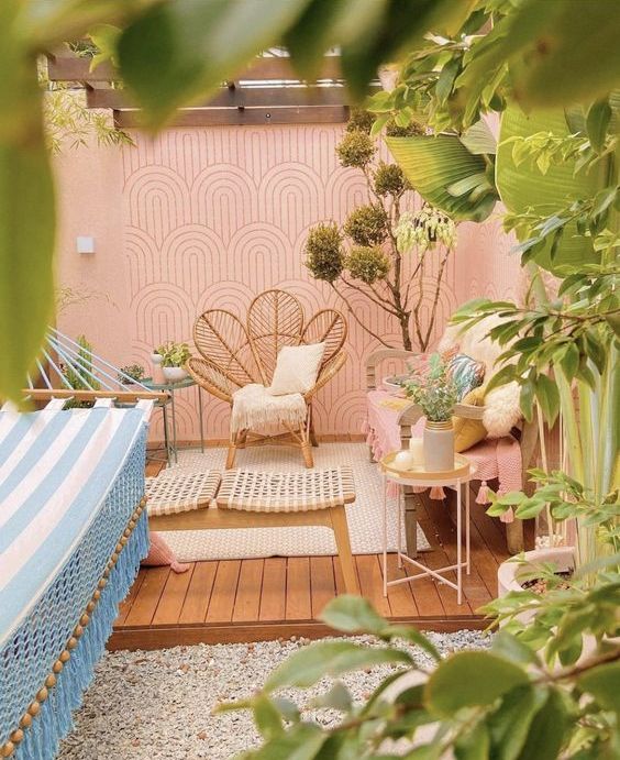 a welcoming patio with pink walls, rattan furniture, a hammock, a bench and lots of plants, blooms and a tree