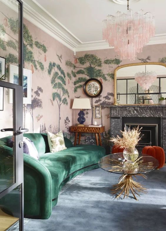 a whimsical living room with pink botanical wallpaper, a marble fireplace, a green curved sofa, rust poufs, a console table with a lamp and a pink chandelier