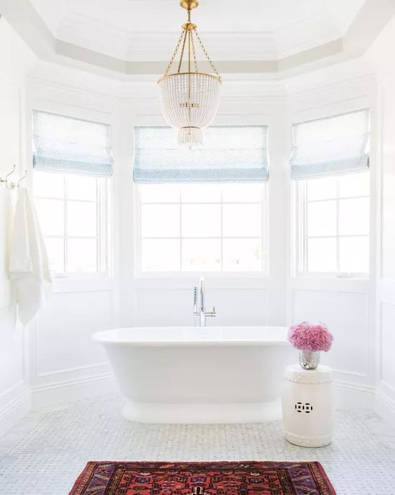 a white bathroom with a bay window, a tub, a crystal chandelier, a side table, a bold boho rug is cool