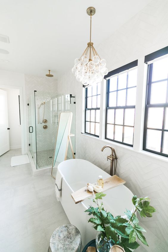 a white farmhouse bathroom with herringbone tiles, a shower, a tub by the windows, a bubble chandelier and some greenery