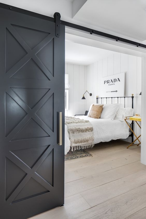 a white farmhouse space with a black barn door, a black metal bed with neutral bedding, a cool folding nightstand and a black sconce