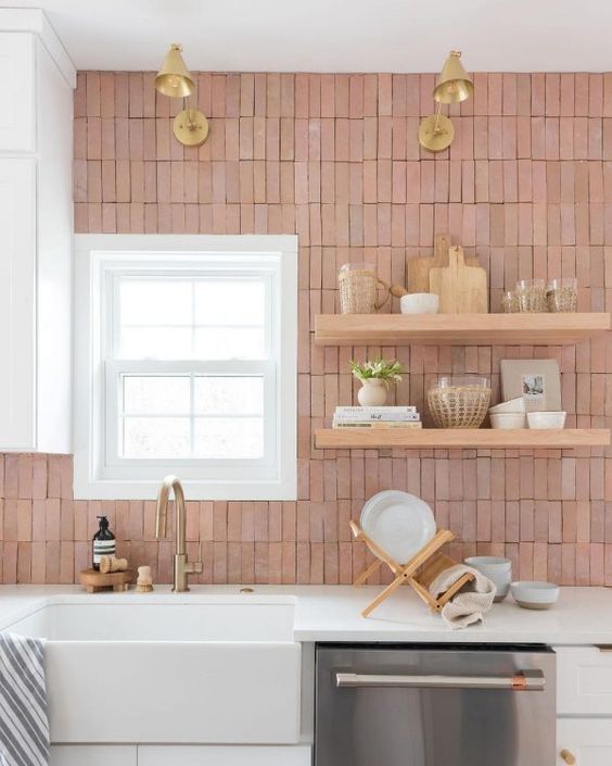 a white kitchen with white countertops, a pink skinny Zellige til backsplash and open shelves, gold lamps and hardware