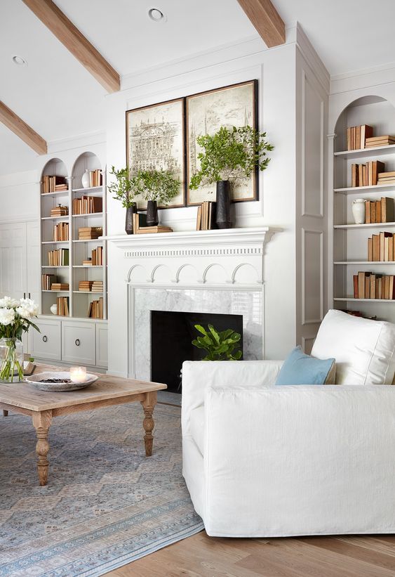 a white living room with a fireplace, arched bookcases, white seating furniture, a whitewashed coffee table and lots of greenery