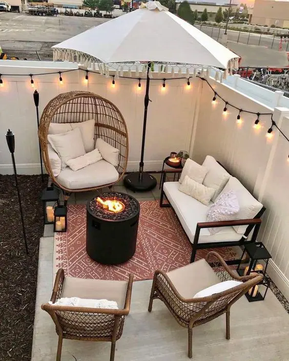 a white patio with a fire bowl, a white sofa and chairs, an umbrella and string lights and lanterns around