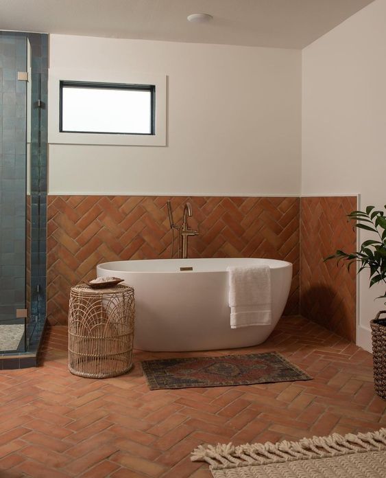 an earthy bathroom clad with terracotta herringbone tiles, a shower done with blue tile, a tub, a side table and a jute rug