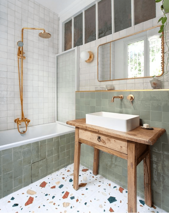 An eye catchy bathroom clad with white and green Zellige tiles, a terrazzo floor, a stained vanity and gold fixtures