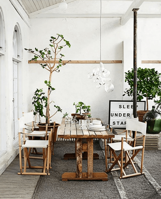 a Nordic summer terrace in neutrals, with wooden and rattan furniture, potted greenery and trees and a bulb chandelier