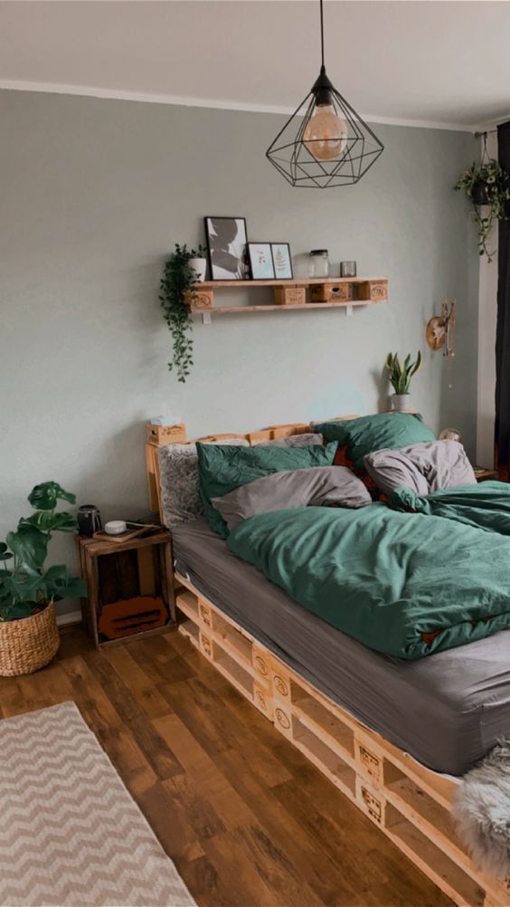 a Scandinavian bedroom with a grey accent wall, a pallet bed with grey and green bedding, a shelf, a pendant lamp and some greenery