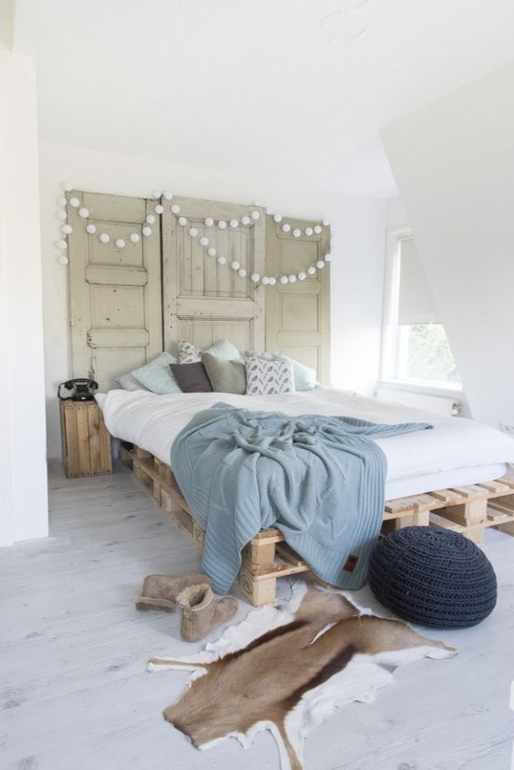 a bedroom with a pallet bed with a neutral bedding, a shutters headboard, a black pouf and some lights over the bed
