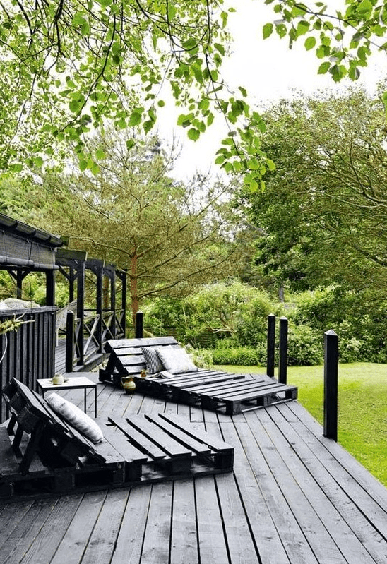a black Scandinavian terrace with pallet loungers, a small table and printed textiles plus lots of greenery around