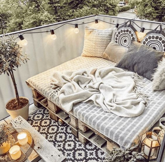 a boho balcony with a pallet loveseat, some boho pillows, a potted plant, candles and lights is amazing