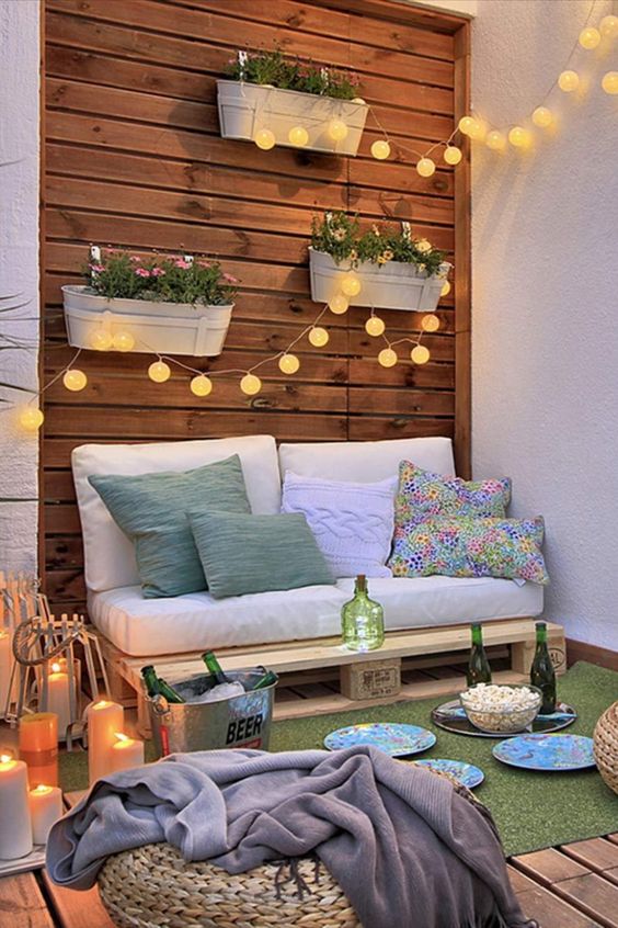 a boho balcony with a small pallet sofa, lights, wall planters, a rug, jute poufs and candles around