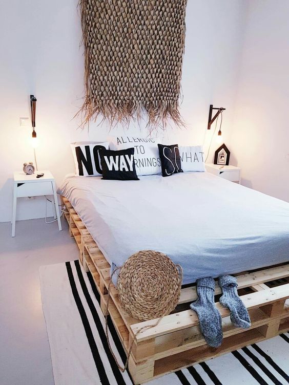 a boho beach bedroom with a pallet with monochromatic bedding, a striped rug, white nightstands, wall sconces and a boho rug