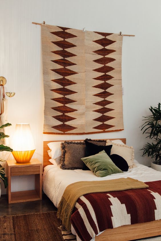 a boho bedroom with a bed and boho bedding, a printed boho rug, a nightstand with a lamp and some potted greenery