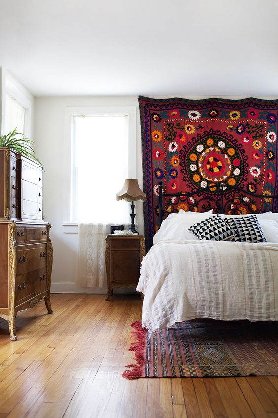 a boho bedroom with a bright rug on the wall, a bed with neutral bedding, vintage stained furniture and a boho rug on the floor