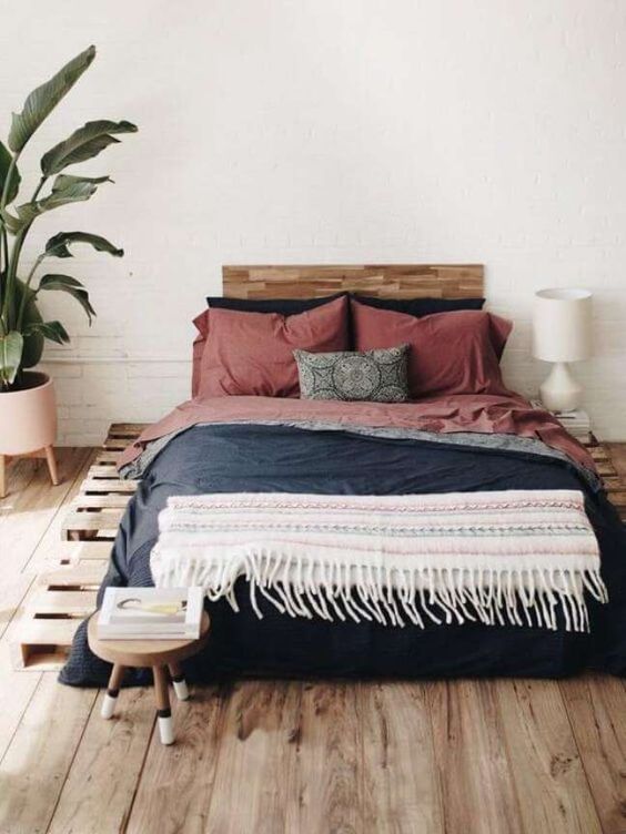 a boho bedroom with a pallet bed and bright bedding, a statement plant, a white lamp and a wooden stool