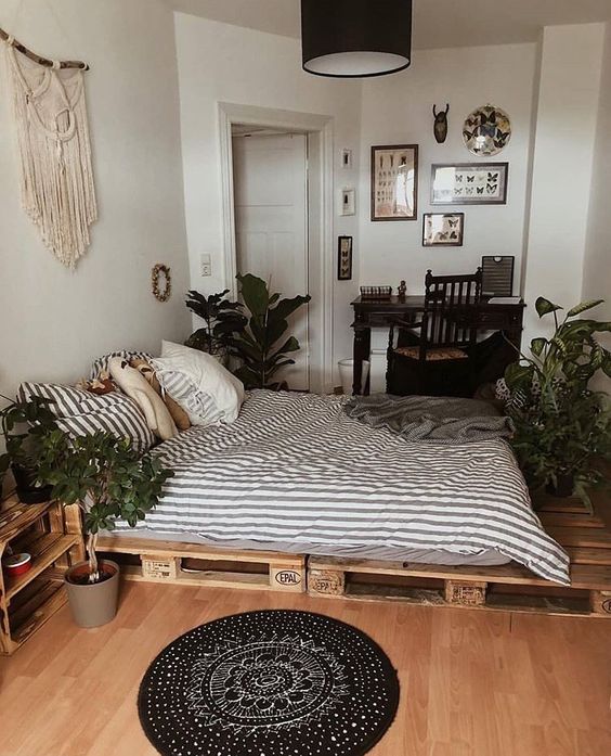 a boho bedroom with a pallet bed and striped bedding, a boho rug, statement plants, boho decor, a black desk and a gallery wall