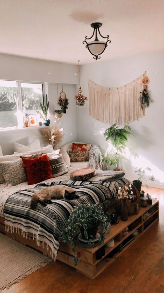 a boho bedroom with a pallet bed with bright bedding and some storage space, fringe on the wall, potted greenery and colorful pillows on the chair