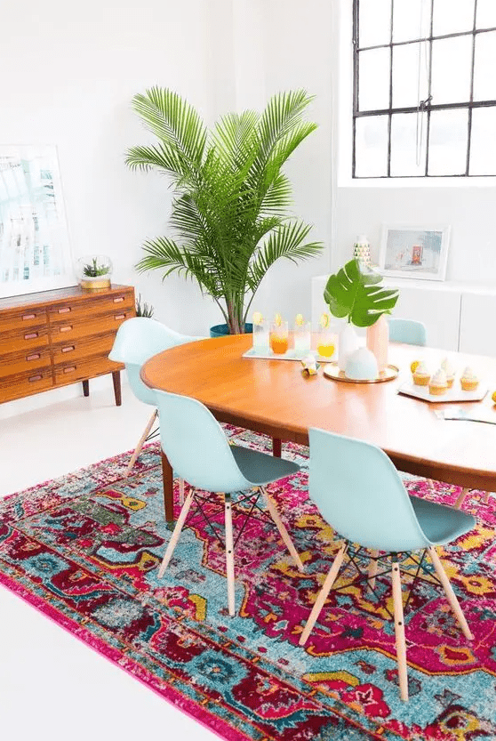a boho dining room with a stained credenza, a stained table and blue chairs, a colorful boho rug and some greenery