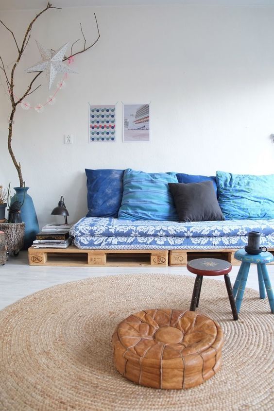 a boho living room with a blue pallet sofa and pillows, stools as tables, a leather pouf and some pretty decor
