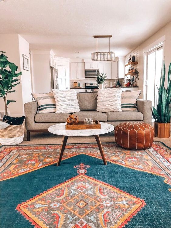 a boho living room with a bold rug, a grey sofa with striped pillows, potted plants, a leather pouf and a coffee table