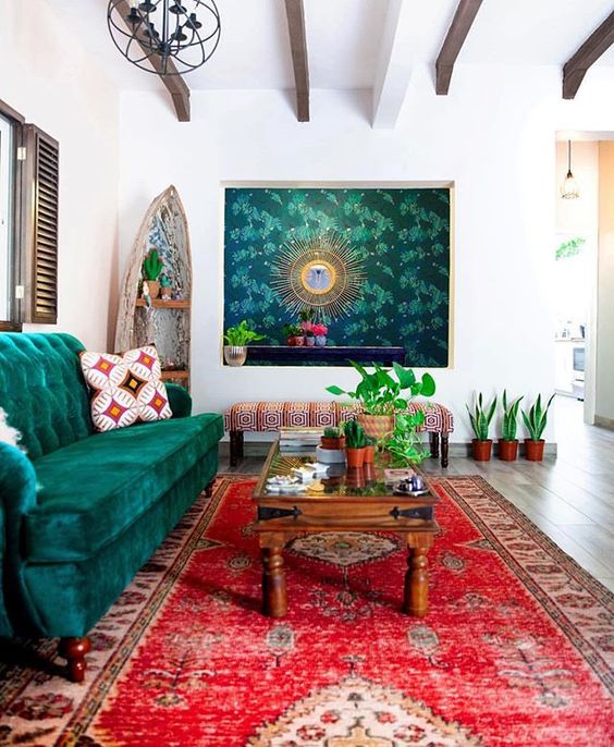 a boho living room with a bright red rug, an emerald sofa, a bold artwork and potted greenery and a stained coffee table