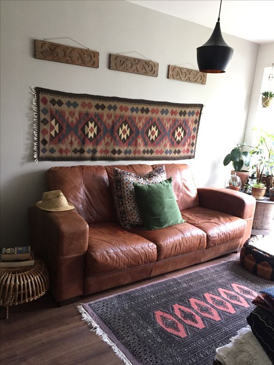 a boho living room with a brown leather sofa and pillows, a boho rug on the wall and floor, a black pendant lamp, a chest with potted greenery