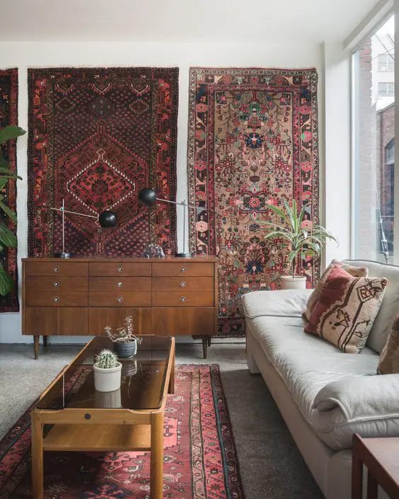 a boho living room with a grey sofa, some stained furniture, a row of boho rugs on the wall and some greenery