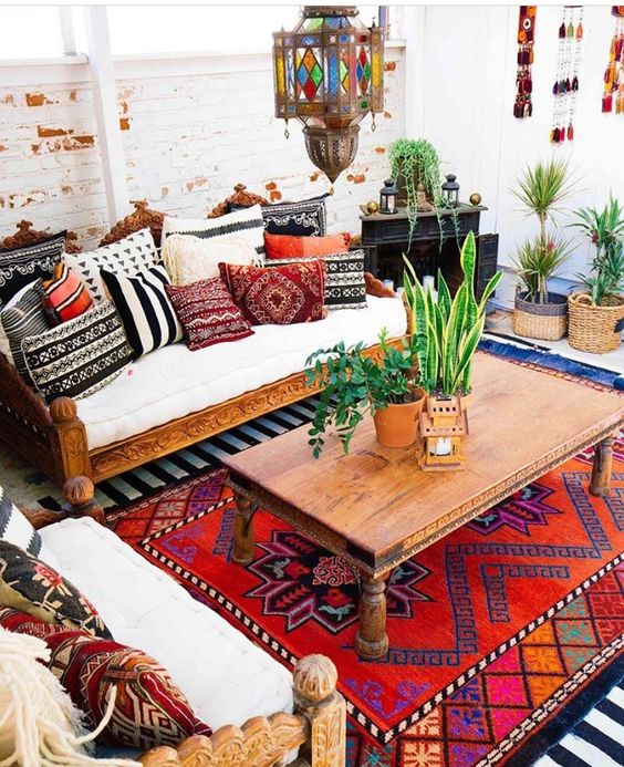 a boho living room with carved wooden furniture and bright pillows, a bold boho rug, a coffee table and potted greenery