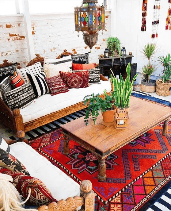 a boho living room with carved wooden furniture and bright pillows, a bold boho rug, a coffee table and potted greenery