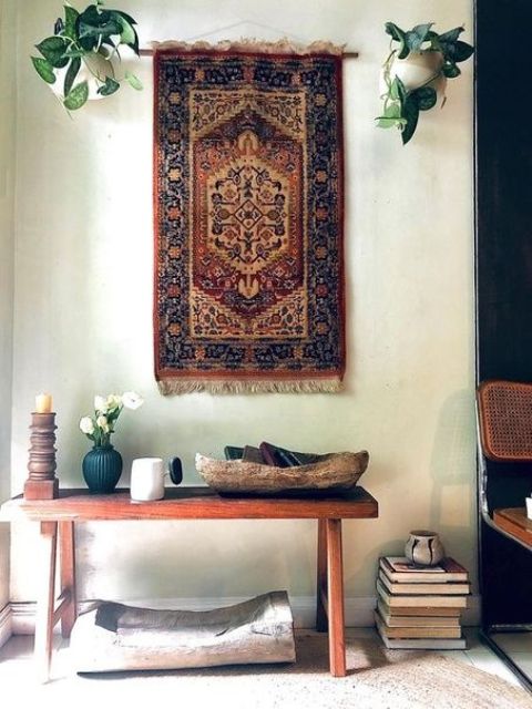 a boho nook with a bench and some decor, a book stacked and candles, a printed boho rug and greenery in pots hanging over the space