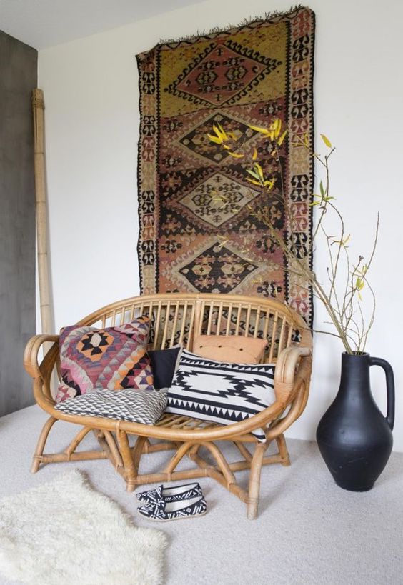 a boho nook with a boho rug on the wall, a rattan loveseat with boho pillows, layered rugs and a vase with branches