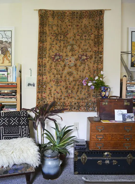 a boho nook with a bright boho rug, a stacke of chests, a boho chair and potted greenery and blooms and some lovely decor