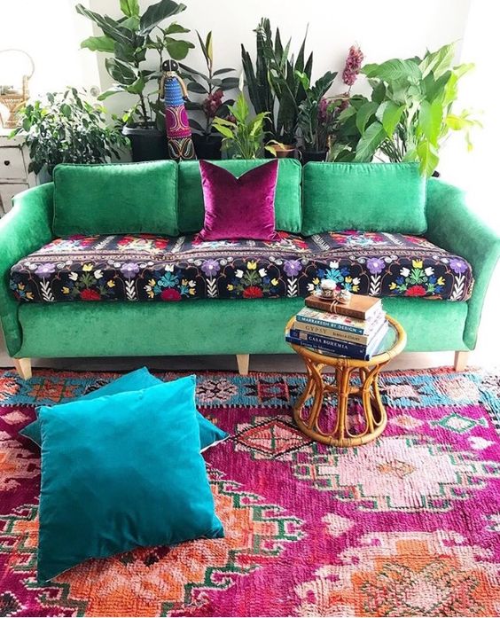 a boho space with an emerald sofa and bold cushion, a colorful rug and navy pillows, a rattan side table and lots of greenery