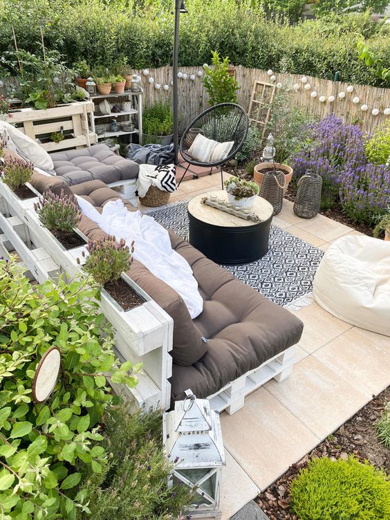 a boho terrace with a pallet corner sofa, a coffee table, a black chair and lots of greenery and blooms around