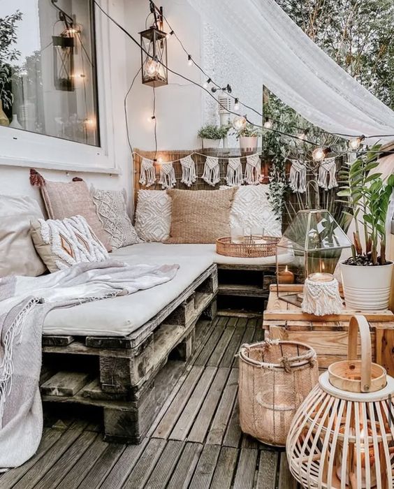 a boho terrace with a pallet sofa and pillows, a macrame banner, a pallet table, lanterns and greenery plus lights around
