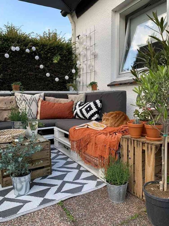 a boho terrace with a white pallet sofa and boho pillows, a crate table and some potted plants, lights and boho blankets